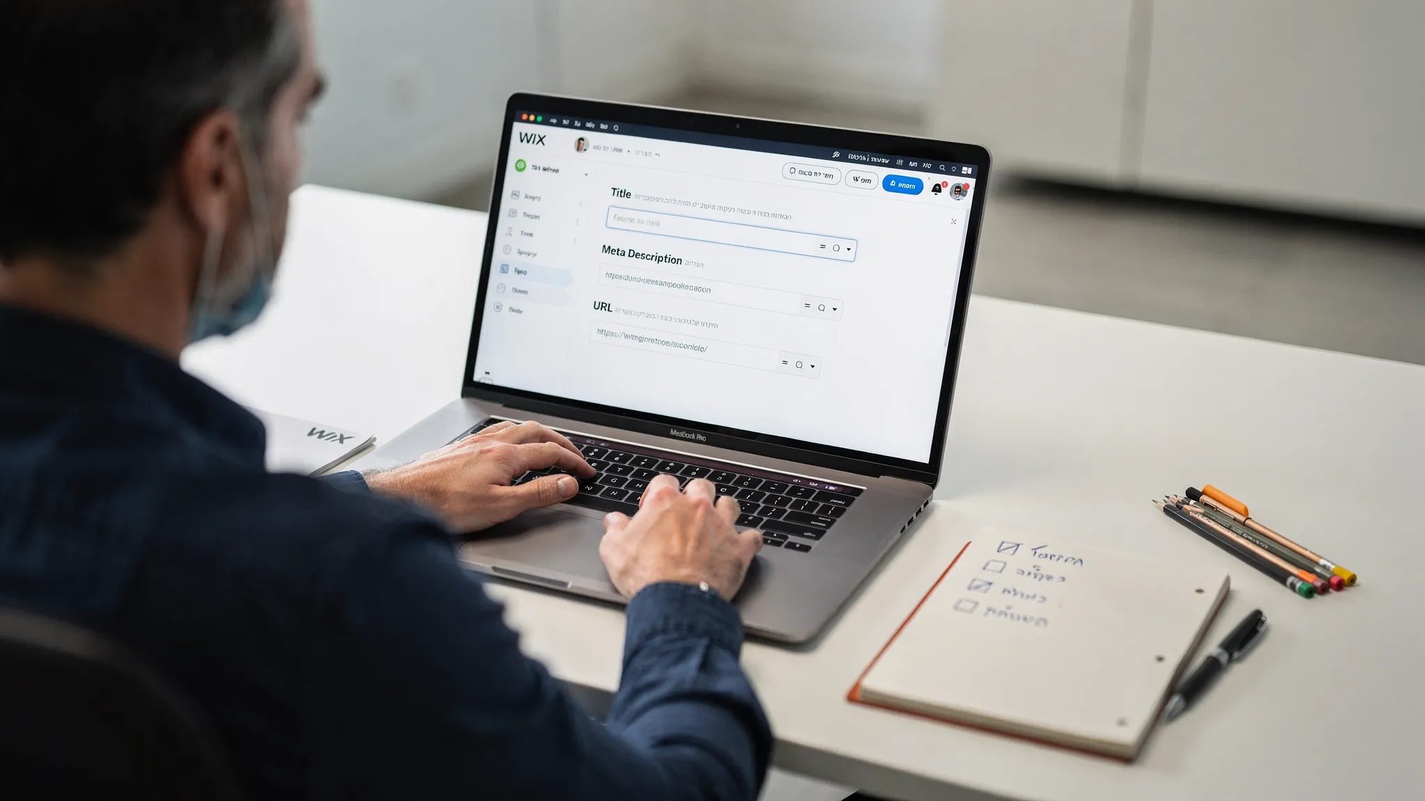 אדם יושב מול לפטופ פתוח שמציג לוח הגדרות SEO של Wix, עם שדות לעריכת Title, Meta Description ו-URL, והמסך פונה לכיוון הצופה. ליד המחשב יש מחברת עם צ׳ק-ליסט קצר וכלי כתיבה.